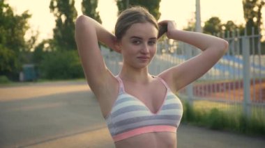 Spor kıyafetli neşeli genç bir kadın gün batımında spor egzersizleri için saçını eline alıyor.