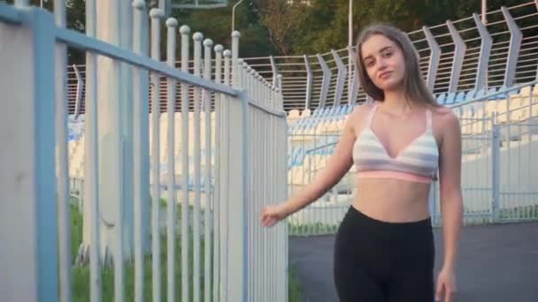 Jolie jeune femme avec de longs cheveux blonds marchant en vêtements de sport le long de la clôture métallique du stade fermé — Video