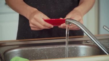 Eller musluğun altında kırmızı tabağı duruluyor. Mutfak lavabosunda bulaşıkları yıkayan tanınmayan biri. Parmaklar akan suyun altında sofra takımını tutuyor. İnsan dikkatli bir şekilde taze akarsu tabakasını temizler