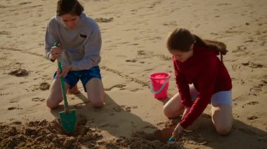 Oğlan büyük kürekle kum kazarken kız küçük kürek kullanıyor, ikisi de gülümsüyor. Erkek ve kız kardeş sahilde kovayla delik açıyor, güneşli günün tadını çıkarıyorlar. Gençler kumsalda birlikte çalışıp neşeli inşaatlar inşa ediyorlar.