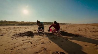 Gençler sıcak günbatımında ıslak kumda büyük çukurlar kazıyorlar. Erkek kardeş yeşil kürekle çalışırken kız kardeş küçük mavi kürek ve pembe kovayla yardım ediyor. Çocuklar boş kumsalda kumlu yapılar yaratıyor, neşeli ve özgür hissediyorlar.
