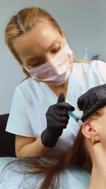 Kozmetik işlem sırasında kulak bölgesine yakın bir yerde dikey oryantasyon uygulayan bir kozmetolog. Estetik uzmanı odaklanmış hassasiyetle yüz gençleştirme için tedavi uyguluyor. Pratisyen hedef alınan cilt geliştirme formülünü enjekte ediyor