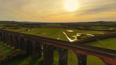Parlak günbatımının altında kırsal kesime uzanan güzel bir viyadük. Gün batımında huzurlu vadinin üzerinde güzel kemerli viyadük. Görkemli tuğla köprü sıcak altın gece ışığı ile yuvarlanan tarlaları kaplıyor