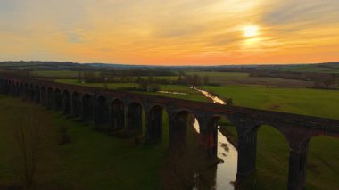 Gün batımında dolambaçlı nehir boyunca uzanan sonsuz demiryolu viyameti. Altın gün batımının sardığı yemyeşil vadideki viyadük. Büyük tarihi köprü, sıcak bir akşamın ışıltısı altında yuvarlanan kırsal bölgeye doğru uzanıyor.