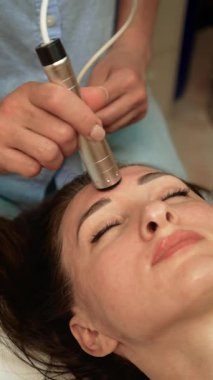 Cilt bakımı uzmanı alnında Biyoelektrik Gençleştirme Terapisi uyguluyor, dikey yönelim. Güzellik uzmanı kolajen üretimini uyarmak için elektrik darbeleri kullanıyor. Mikro akım kaldırma ile cilt esnekliğini geliştiren estetik uzmanı