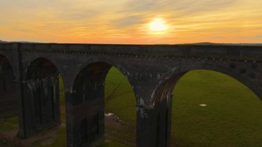 Gün batımında gökyüzüne doğru yükselen tuğla viyadük kemerlerinin yakın görüntüsü. Muhteşem İngiliz viyadük kanalı güneşin son ışınlarını yansıtıyor. Tarihsel demiryolu yapısı akşam vakti muazzam yeşil alanlar üzerinde yükseliyor.