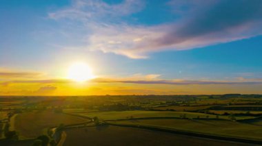 İnsansız hava aracından elde edilen tarım arazisi üzerinde parlayan güneşle gün batımı hızlanıyor. Akşamları, dramatik turuncu gökyüzü ve yuvarlanan tarım arazisi hava manzaralı. Parlak güneş ışığı ve dağılmış bulutlarla altın saat