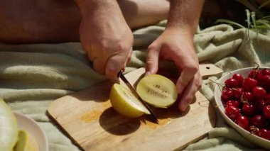 Tahta kesme tahtasında kivi dilimleyen adam piknik için taze meyve hazırlıyor. Erkek eller sulu kiviyi dikkatlice ikiye böler. Olgun kiviyi aile eğlencesi için bölen bir adamla açık hava yemek sahnesi