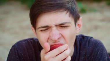 Genç bir delikanlı kırmızı elma yiyor, yoğun bir ifadeyle sulu ve keskin tadını çıkarıyor. Erkek, kırmızı elmayı ısırıyor, odaklanmış enerjisiyle tatlı ve çıtır çıtır tadını çıkarıyor. Kırmızı elmadan hoşlanan adam, ferahlatıcı ve doğal duygulara dalmış.