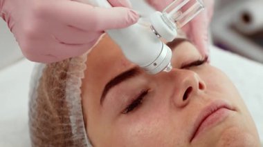 Deri stüdyosunda gelişmiş bir aletle hidro yüz tedavisi gören kadın rahat bir ifadeyle yatıyor. Cilt bakımına giren bir kadın modern ekipman kullanıyor ve sakin görünüyor. Hidro aletle yüz terapisi gören bir kadın, dinginliği gösteriyor.
