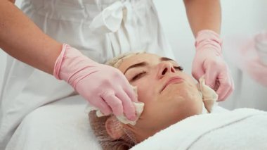 Kozmetik kliniğinde güzellik ameliyatı geçiren bir kadın yüz bakımı sırasında rahat bir şekilde uzanıyor. Güzellik uzmanı aşırı ürünü çıkarmak için kadın yüzünü nazikçe siliyor. Profesyonel cilt bakımı yapan hasta yenilenmeye odaklanmış durumda.