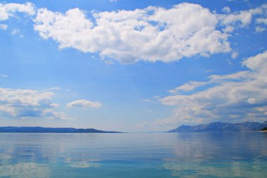 Güzel mavi gökyüzü, kumsaldaki sakin denizin üzerinde beyaz bulutlar.