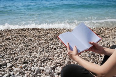 Genç güzel bir kız sahilde deniz kenarında kitap okuyor.