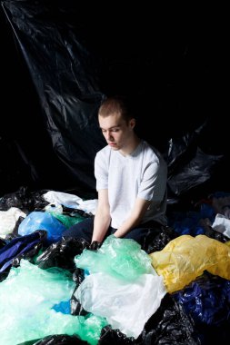 Plastik torba çöpüne oturan bir adam, çevresel bilinç konsepti, stüdyoda çekim yapmak, karanlık ışık arka planı..