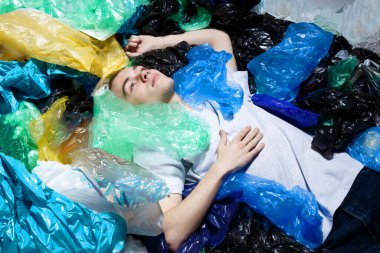 Mavi gözlü genç adam, uzanmış, burnunda halkayla, etrafı plastik torbalarla çevrili, yukarı bakıyor. Yatay görünüm.