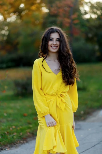 Portrait of a beautiful smiling teen girl with brown wavy hair, wear in yellow dress, posing in a park background. — Stock Photo, Image