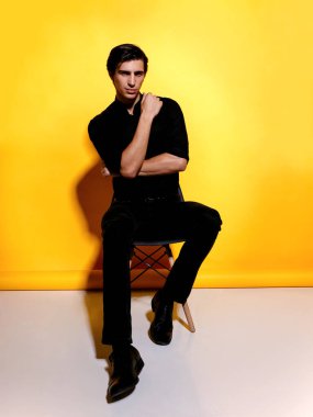 Sarı arka planda oturan, siyah elbiseli, neşeli, yakışıklı genç bir adamın portresi. Dikey görünüm.