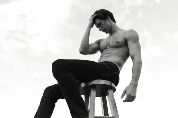 Hezký svalnatý muž bez košile pózující venku. Fotka bílá a černá. Mužský model sedící na židli. — Stock fotografie