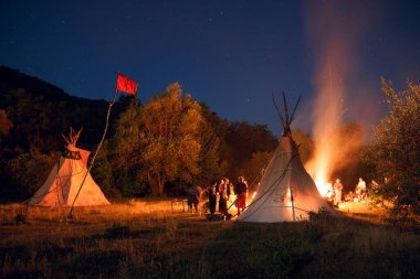 Karanlık orman arka planında çadır ve büyük şenlik ateşi olan gece ormanlarında insanlarla kamp yapmak. Yatay dış çekim.