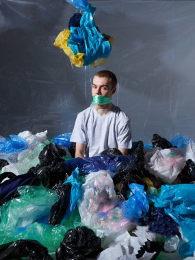 Dikey görünüm. Genç adam, plastik torbayla ağzına bağlanmış, plastik poşetlerle çevrili, kameraya bakıyor..