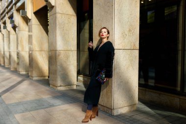 Yatay görüş. Şık elbiseli ve lüks montlu muhteşem şehvetli bir kadının şehir önünden geçerken çekilmiş moda fotoğrafı..