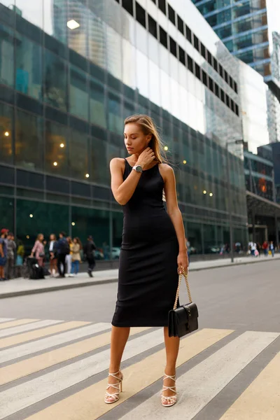 Full-length frontal portrait of fashionable girl in the city. She walks on the crosswalk. Wearing in black slim dress and white high heels, holding in hand a black bag.