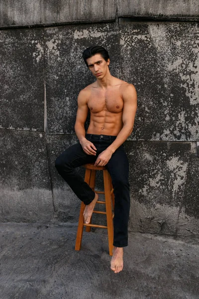 高い椅子に座って 古い黒い壁の近くに裸の胴を持つ魅力的な若い男の完全な長さの肖像画 健康な男だ — ストック写真