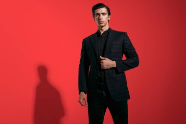 Yatay görüş. Kırmızı arka planda siyah takım elbiseli, kendine güvenen esmer bir genç..