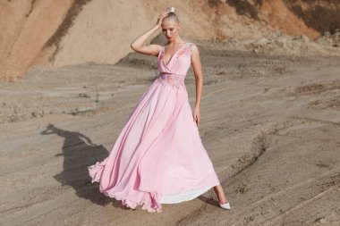 Pembe uzun elbiseli sarışın bir kızın uzun boylu portresi güneşli bir yaz gününde kumda poz veriyor. Yatay görünüm.