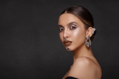 Alçak kesim siyah elbiseli, makyajlı, gümüş bijouterili, gri arkaplandan kameraya bakan şehvetli bir kadının profili. Yatay görünüm.