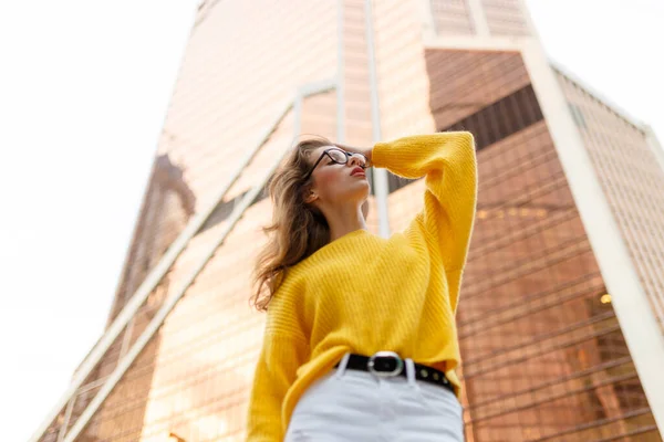 水平方向の眺め 波状の髪をした若い女性の肖像画 現代的な建物の後ろに黄色のセーターと白いジーンズを着て — ストック写真