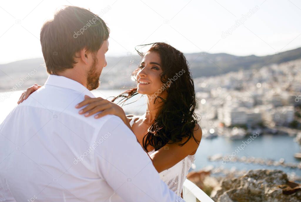 Rear view of a beautiful just married couple, wearing in white clothing, with back panorama of city, wedding in Greece.