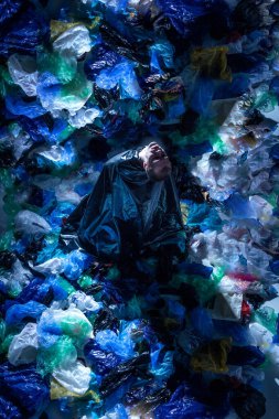 Stüdyodaki boş plastik çöp torbalarıyla çevrili genç bir adamın sanatsal görüntüsü. Ekolojik sorunlardan memnun, kirliliğin kanıtı, stüdyonun karanlık duvarında izole edilmiş..