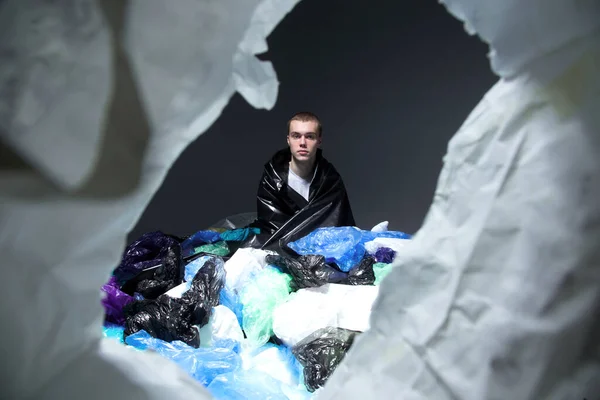 Frontal portrait of a young man with haircut seated and covered with plastic bag garbage, dark light background.