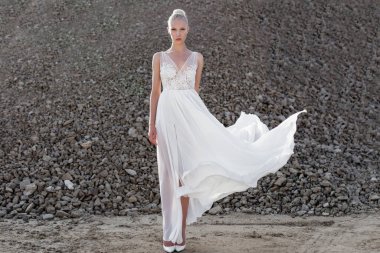 Yatay görüş. Beyaz gelinlik giymiş, rüzgarda uçan, kameraya bakan, gün boyunca taş yığınlarının arkasında poz veren sarışın genç kadının ön görüntüsü..