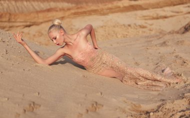 Yatay görüş. İnce, pahalı elbisesi olan sarışın bir kadının çölde, gün batımında kumların üzerinde poz vermesi..