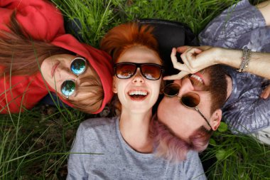 Gülümseyen, güneş gözlüklü, yeşil çimenlere uzanan, güneş gözlüğü takan ve kameraya bakan gençlerin en güzel manzarası..