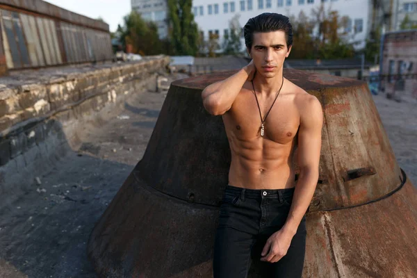 Frontal portrait of a athletic young man with naked torso posing on the roof at the sunset background.