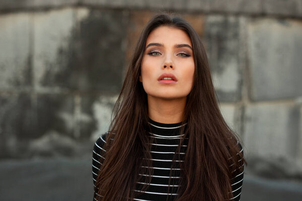 Frontal portrait of a beautiful girl with straight brunette hair and makeup, looking confident at camera, over black old wall background.