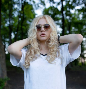 Beyaz bluz giyen, yuvarlak güneş gözlüğü takan, ağaçların arkasından kameraya bakan güzel, kıvırcık, sarışın bir kadının ön portresi..