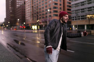 Genç yakışıklı, mutlu hippi genç bir gezgin şehirde yolda otostop çekiyor. Yatay görüş. Metin için boşluk.