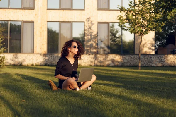 Retrato de una bonita mujer joven con gafas de sol sentada sobre hierba verde con las piernas cruzadas durante el uso de la computadora portátil sonriendo . — Foto de Stock