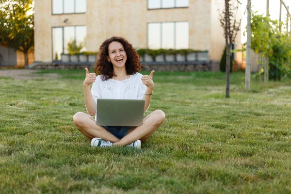 Frontalansicht einer fröhlichen Frau, die mit überkreuzten Beinen auf grünem Gras sitzt, während sie lächelnd ihren Laptop benutzt und die Daumen nach oben hält. — Stockfoto