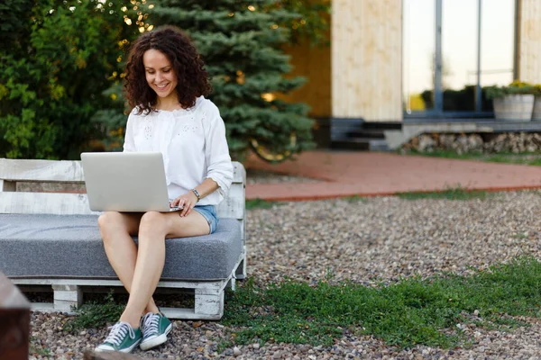 Lächelnde junge Frau in lässiger Kleidung, die zu Hause am Laptop arbeitet, über Hinterhof und häuslichen Hintergrund — Stockfoto