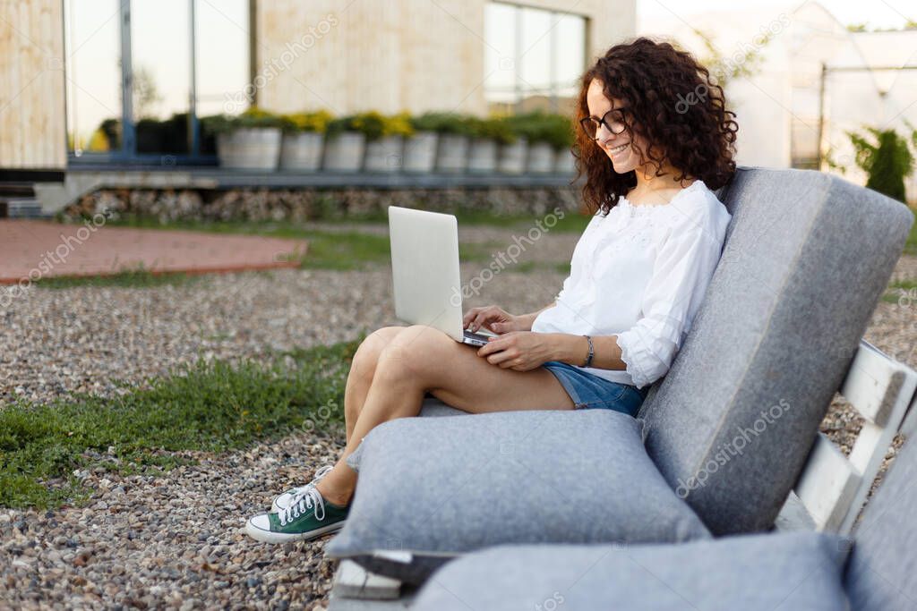 Cheerful young woman dressed in casual clothes, working at home on a laptop, over backyard and home background