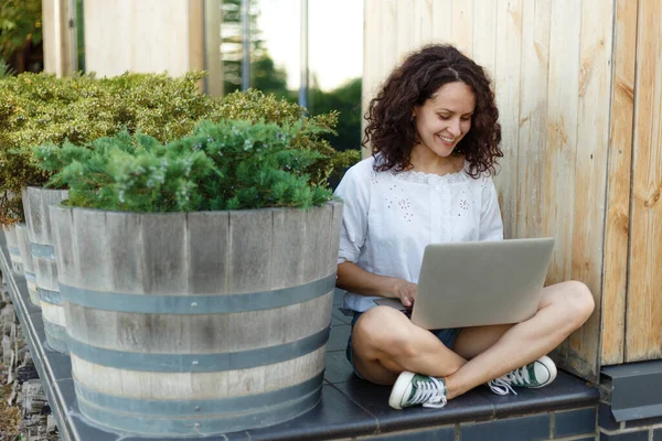 Porträt einer hübschen, lockigen Frau, die an Sommertagen mit überkreuzten Beinen am Laptop sitzt und lächelt. — Stockfoto