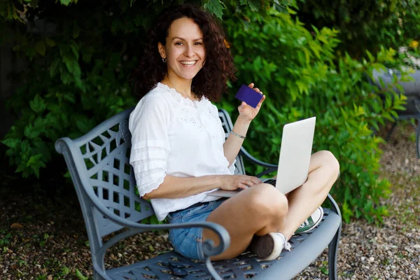 Ein Mädchen, das im Sommer mit überkreuzten Beinen im Park auf der Bank sitzt, kauft online mit Laptop und Kreditkarte. — Stockfoto