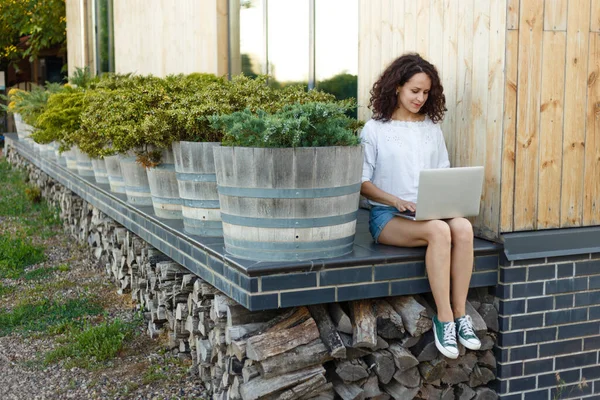 Retrato de una joven bastante rizada sentada afuera durante el día de verano, trabajando en el portátil. Contenido freelancer . — Foto de Stock