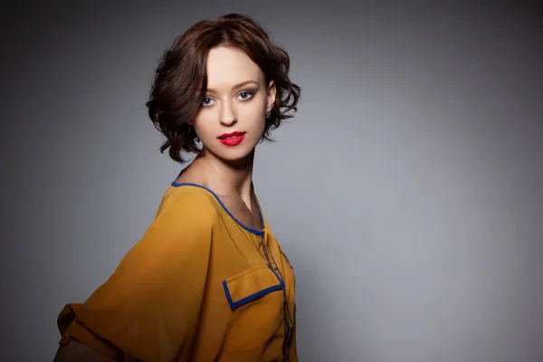 Frontal portrait of a girl with short-haired looking at the camera, with make-up and red lips, wear in blouse of mustard color on a grey background.