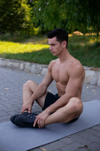 公園でスポーツのためのマットレスの上でストレッチ演習を行うスポーツウェアの裸の胴を持つ若い選手 — ストック写真